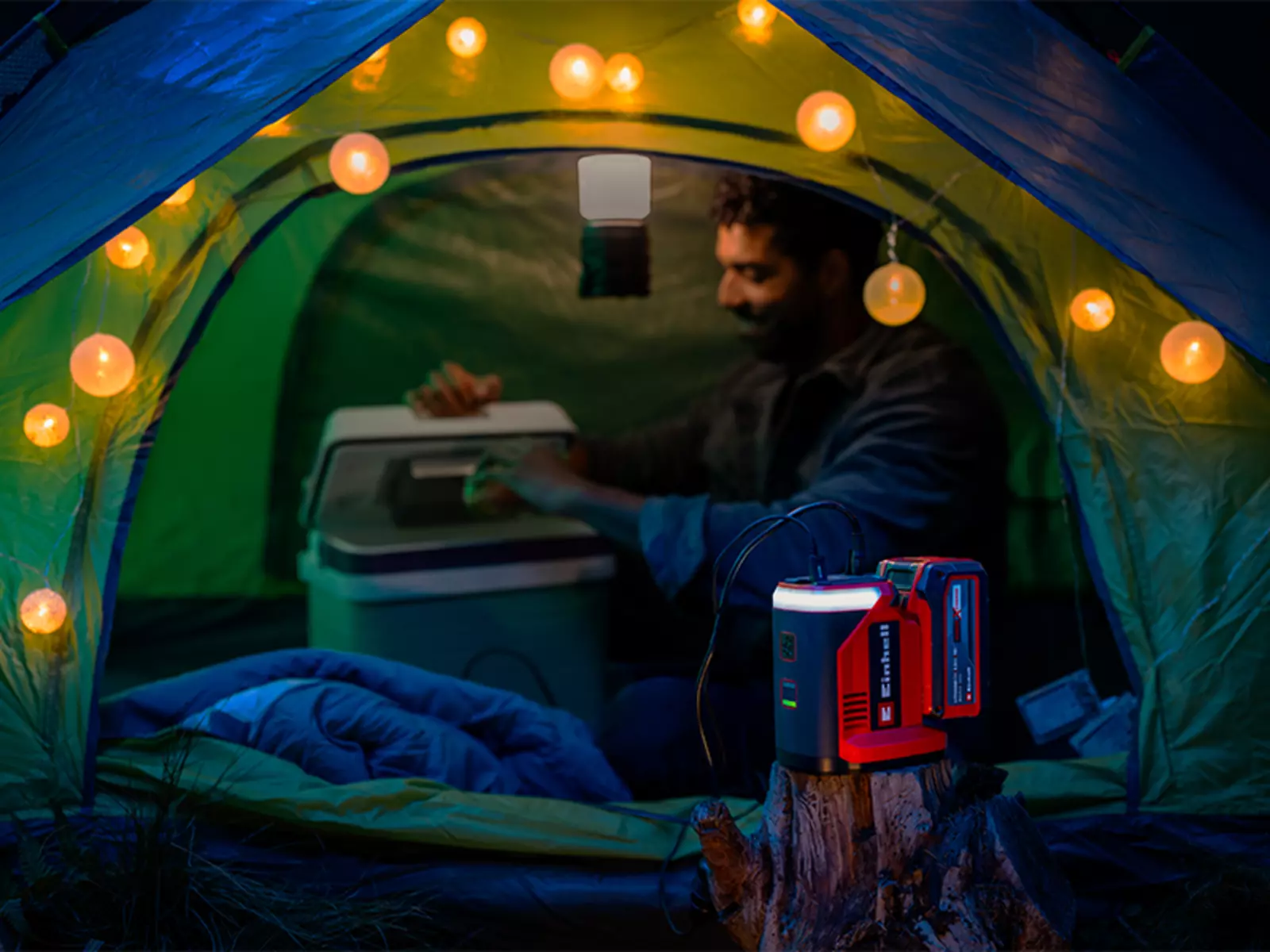 Аккумуляторная электростанция Einhell TE-ES 18/150/1 Li-C-Solo обеспечивает электропитанием гирлянду фонарей во время кемпинга в палатке; на заднем плане мужчина сидит перед открытым холодильником.