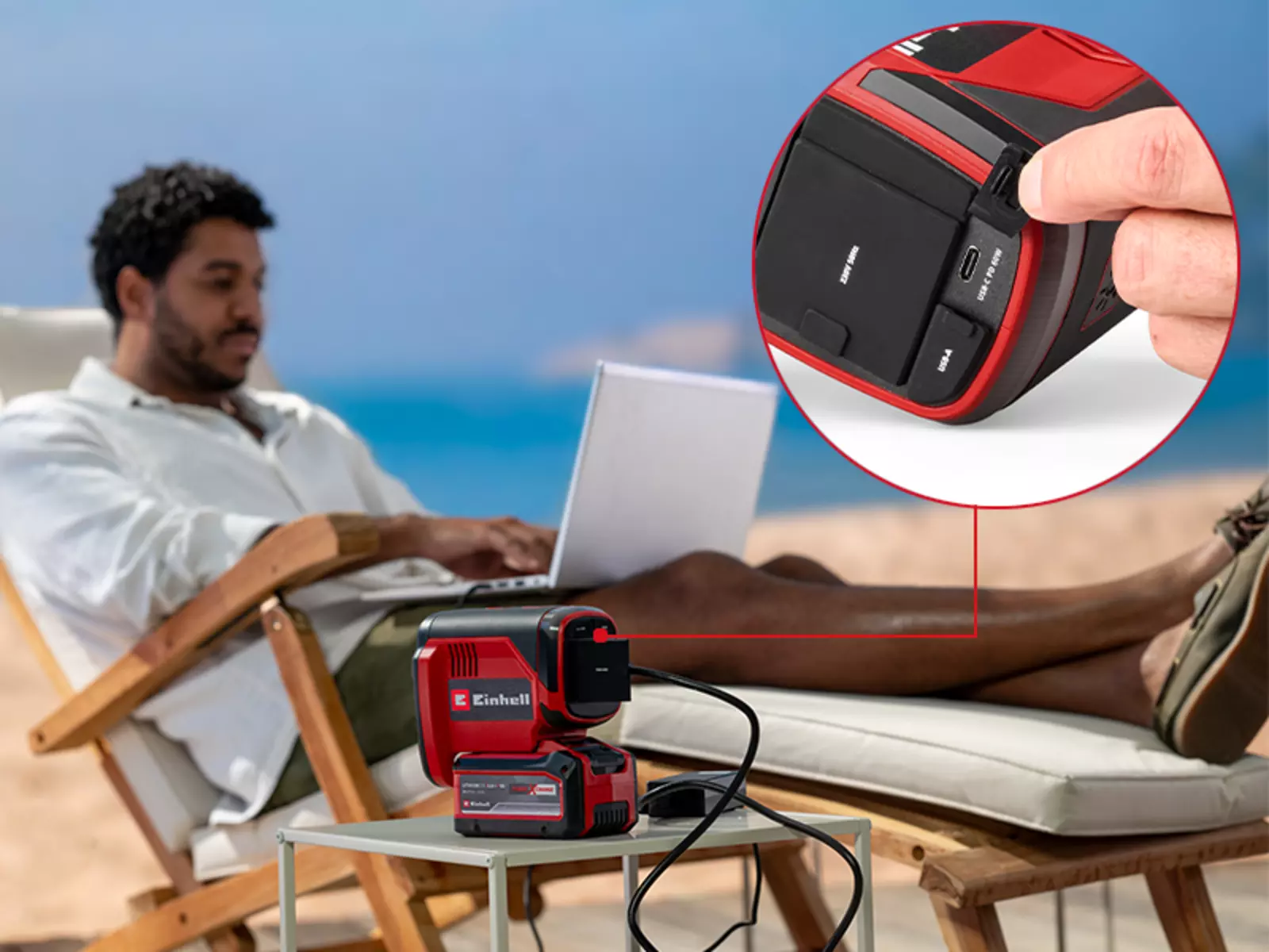 Мужчина сидит на пляже с ноутбуком, работающим от аккумуляторной электростанции Einhell TC-ES 18/150/1 Li-Solo; на крупном плане виден палец, указывающий на порт USB-C электростанции.