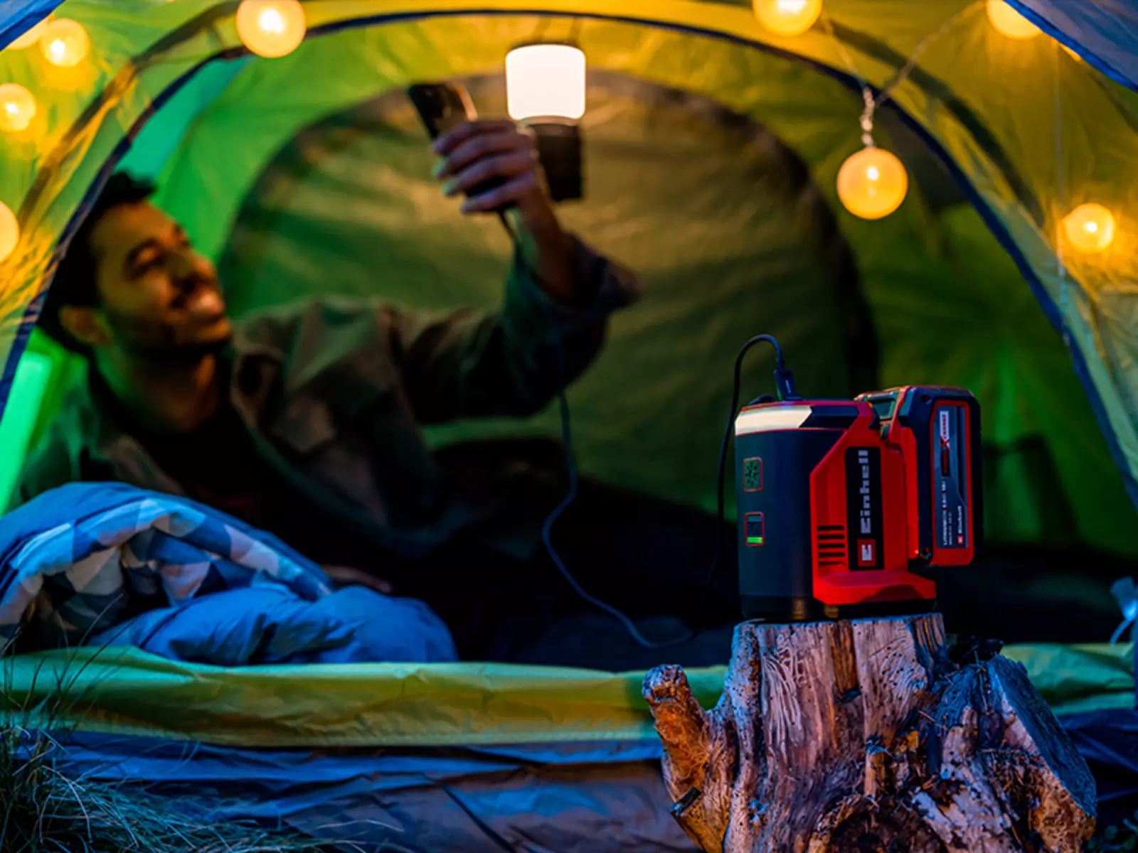 Аккумуляторная электростанция Einhell TC-ES 18/150/1 Li-Solo стоит на пне перед освещенной палаткой, на заднем плане — человек.